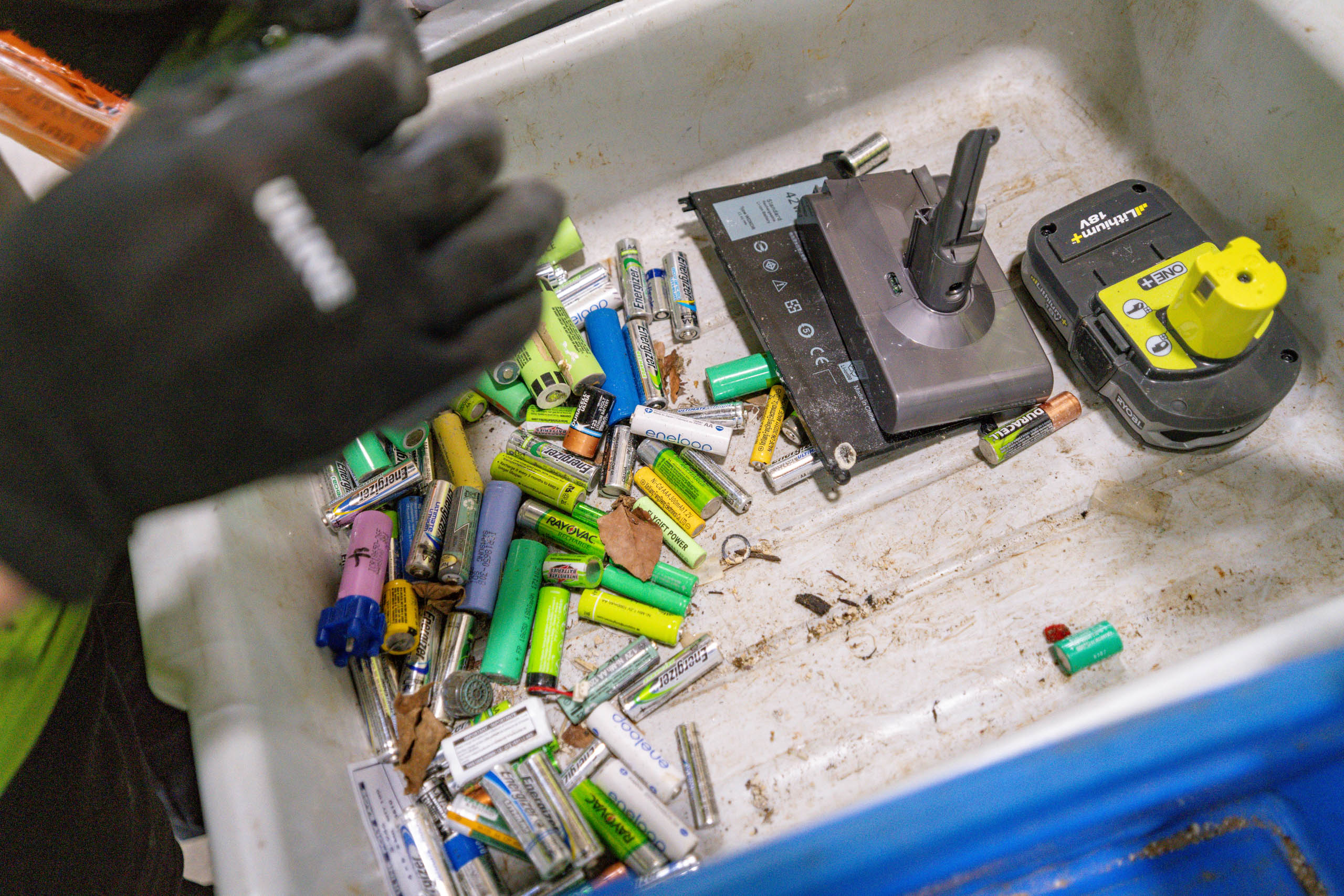 A person wearing black gloves sorts a variety of used batteries and battery packs in a white bin, including AA, AAA, and rechargeable batteries of different colors and sizes.