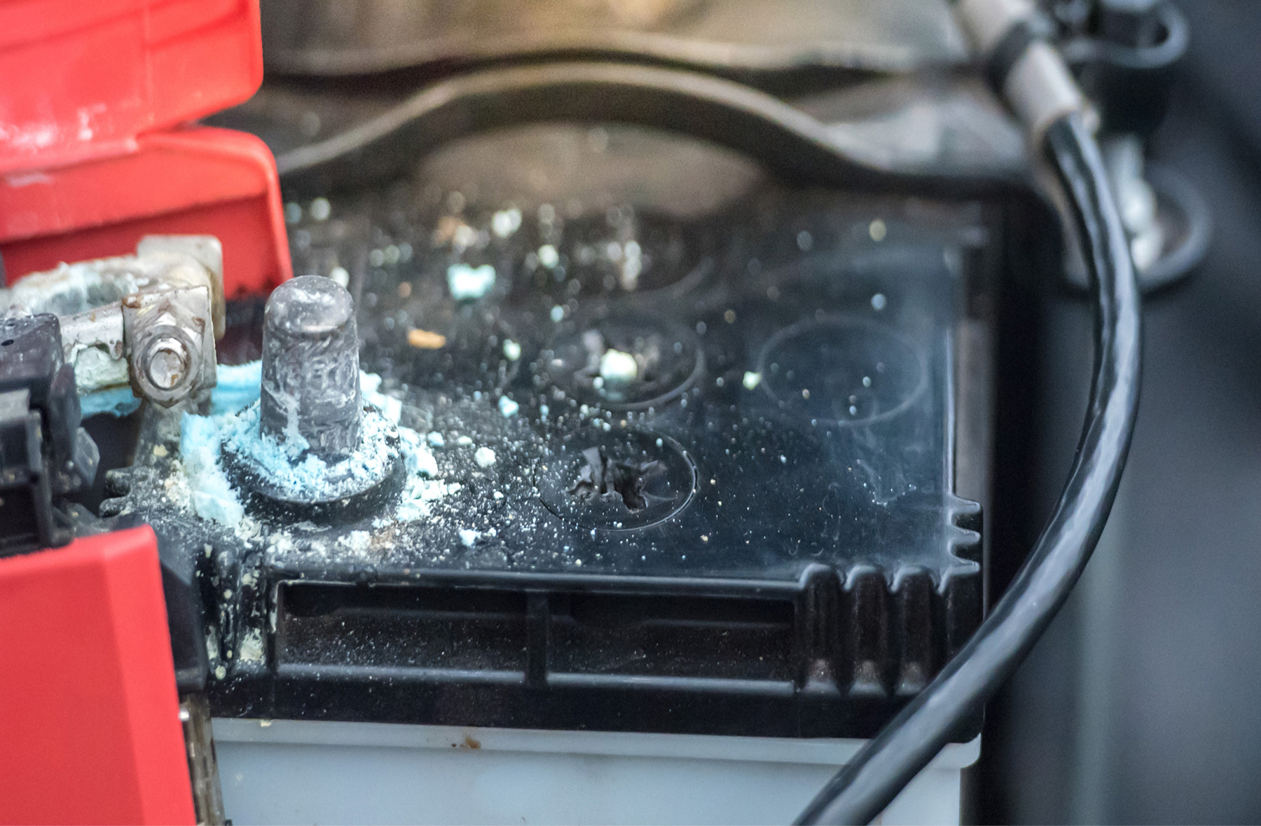 Close-up of a car battery with visible corrosion and white powdery buildup around the metal terminal, indicating possible battery acid leakage or damage. Black cables and red plastic components are also visible.