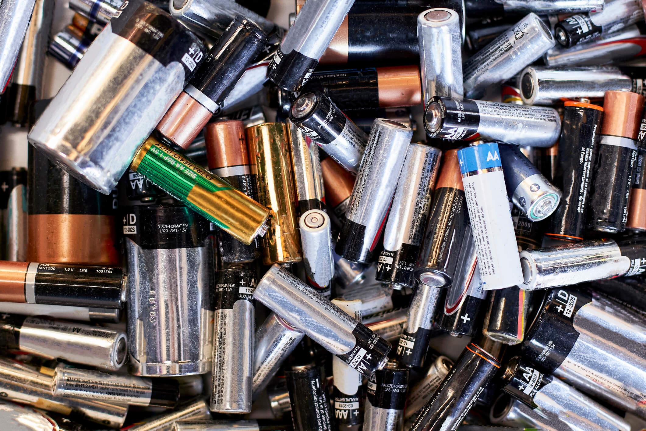 A large pile of used batteries of various sizes and brands, including AA, AAA, and D types, with some showing worn labels and metallic surfaces.